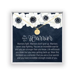 May include: A gold-tone necklace with a small circular pendant featuring three upward-pointing chevrons. The necklace is displayed on a card with white floral illustrations and text that reads: The heart of a Warrior. Warriors fight. Warriors don't give up.