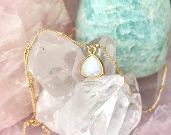 May include: A gold chain necklace featuring a triangular pendant with an iridescent white stone. The pendant is suspended from a delicate gold link chain that includes small gold beads spaced along the length of the chain, creating a refined look.