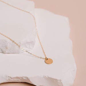 A delicate gold necklace with a small round pendant rests on a white stone surface against a soft pink background.