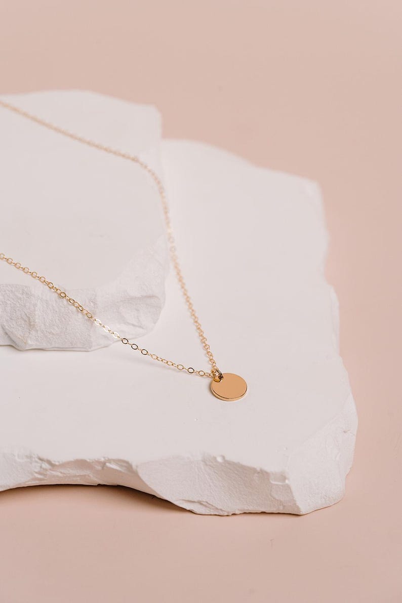 A delicate gold necklace with a small round pendant rests on a white stone surface against a soft pink background.