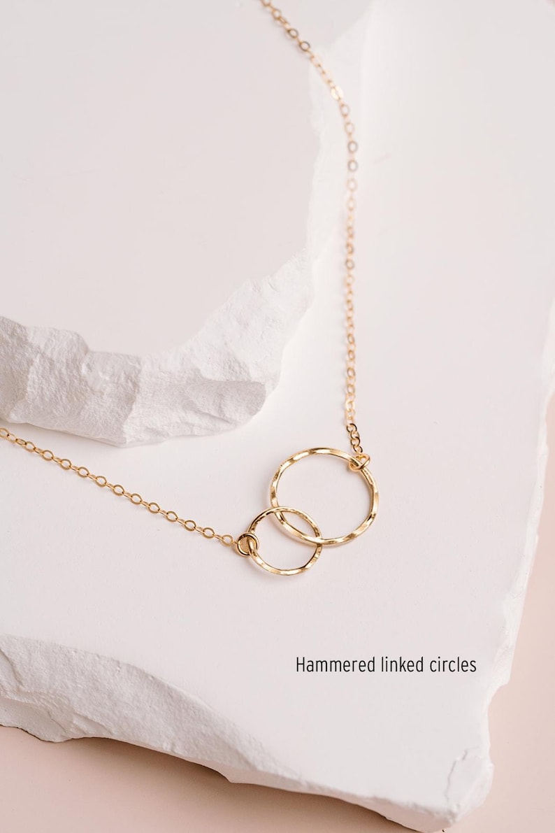 A delicate gold necklace with two intertwined rings, resting on a white stone surface.