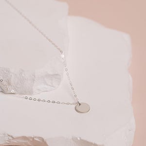 A delicate silver necklace with a round pendant rests on a white stone surface against a soft pink background.