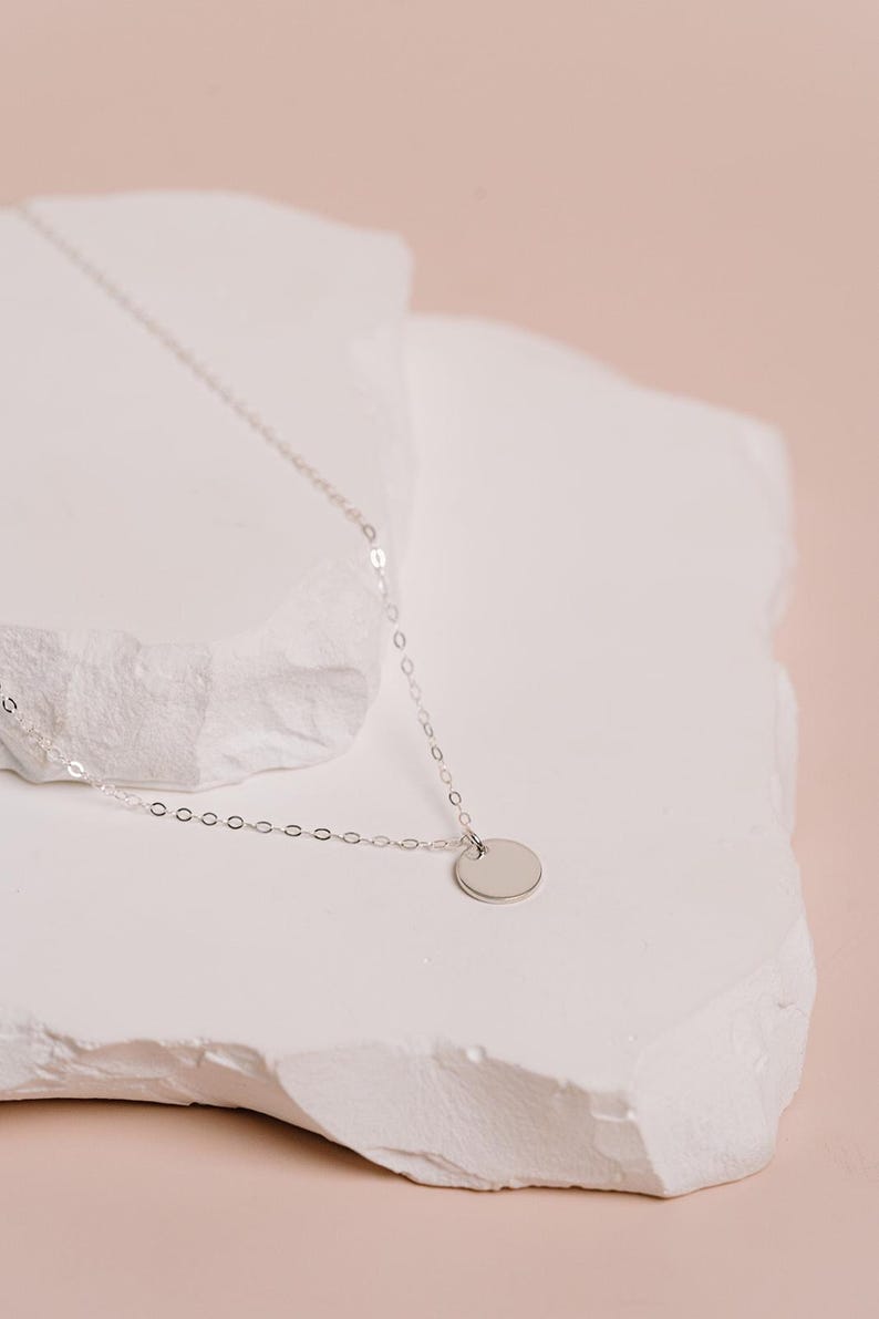 A delicate silver necklace with a round pendant rests on a white stone surface against a soft pink background.