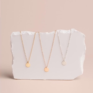 Three delicate necklaces with small circular pendants are displayed on a white stone or marble surface against a light pink background.