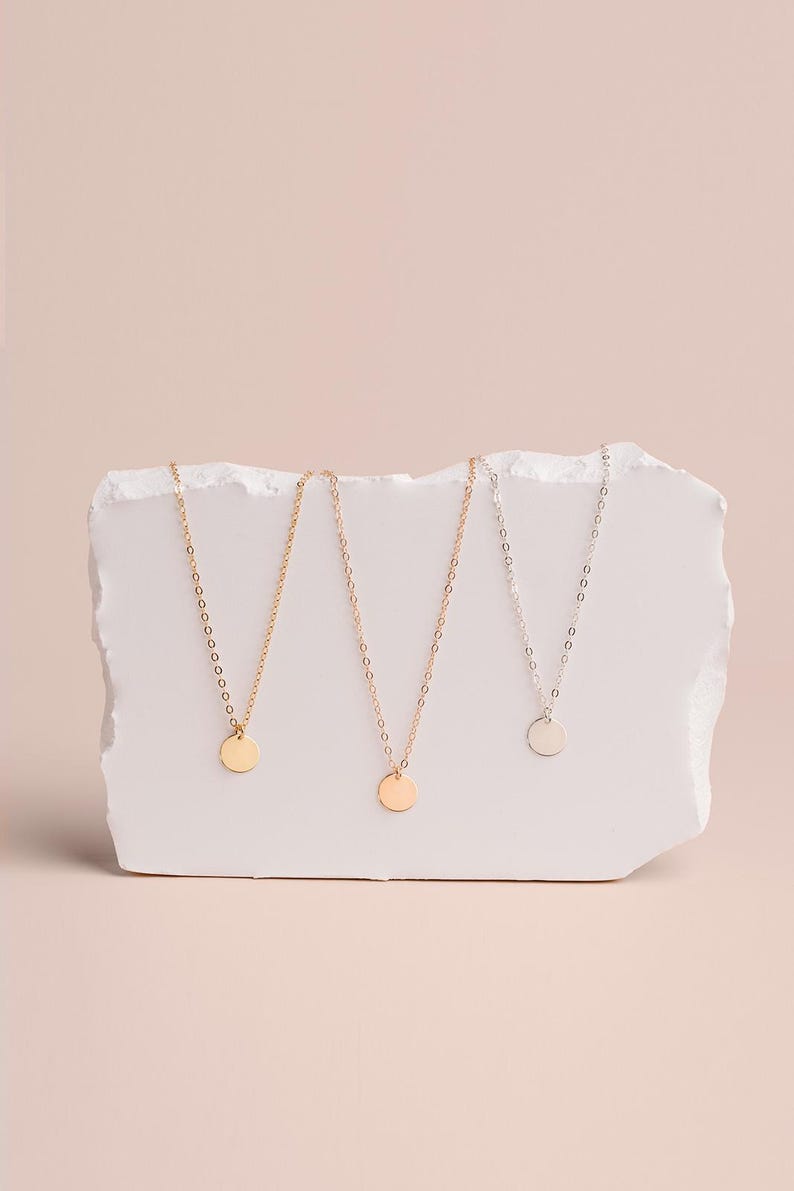 Three delicate necklaces with small circular pendants are displayed on a white stone or marble surface against a light pink background.