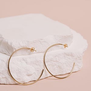 May include: A pair of thin gold hoop earrings with a circular design and post back closures. The earrings feature a smooth, polished metallic finish and a simple, open-ended hoop structure that forms a complete circle when worn.