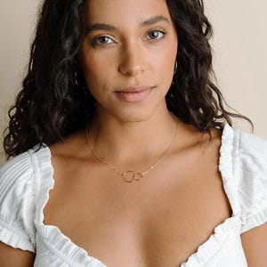 A young woman with dark curly hair wearing a white top and a necklace with a circular design.