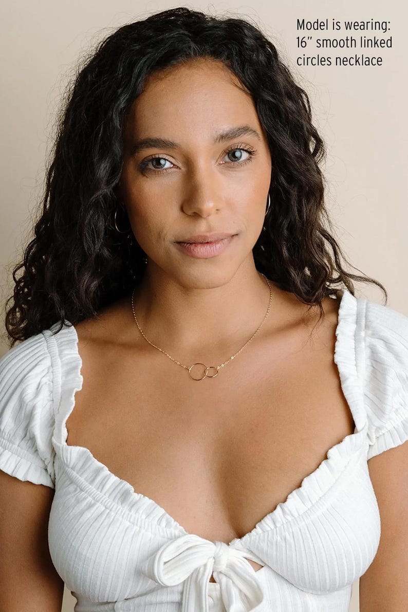 A young woman with dark curly hair wearing a white top and a necklace with a circular design.