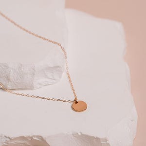 A delicate gold necklace with a round pendant rests on a white stone surface against a peach-colored background.