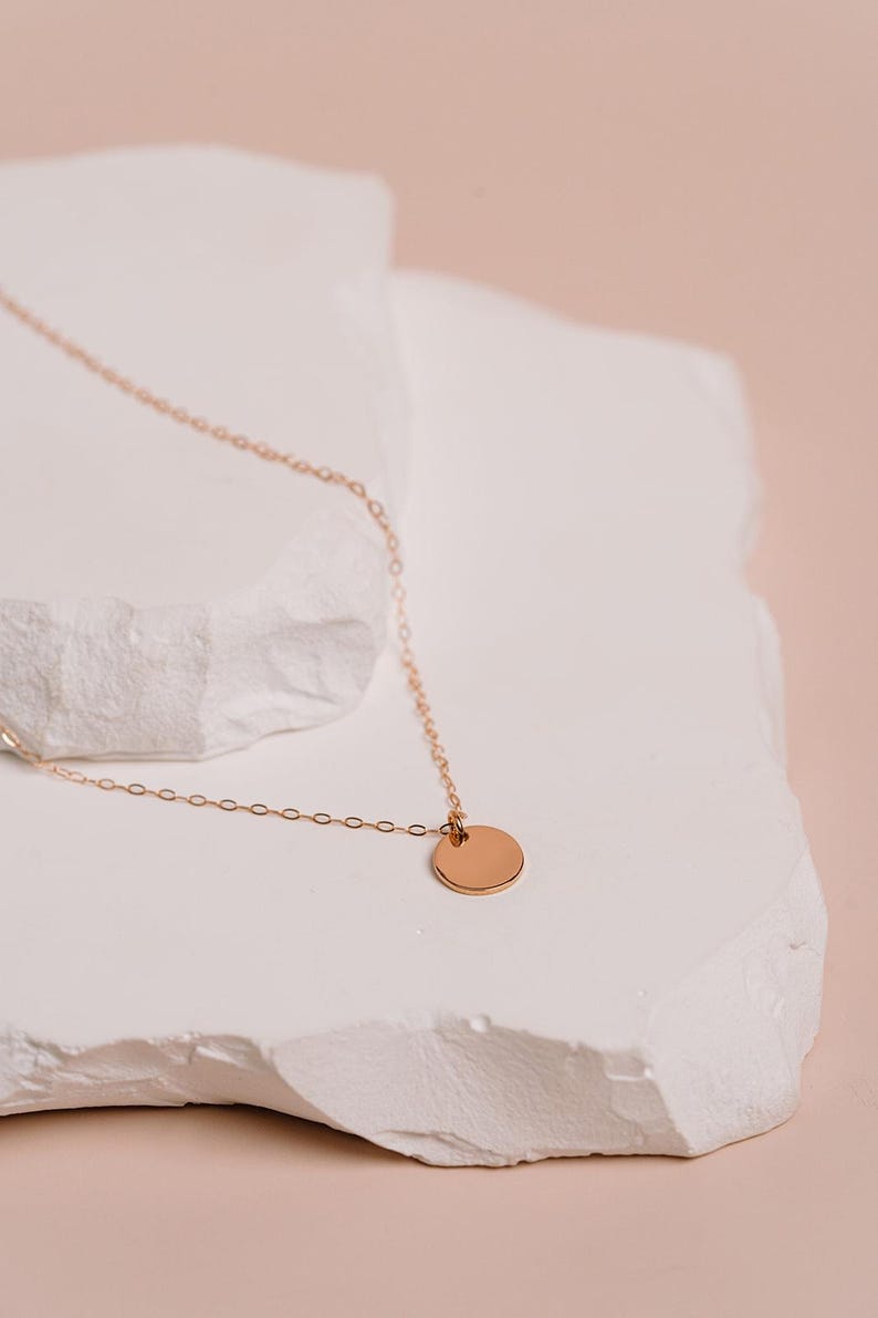 A delicate gold necklace with a round pendant rests on a white stone surface against a peach-colored background.