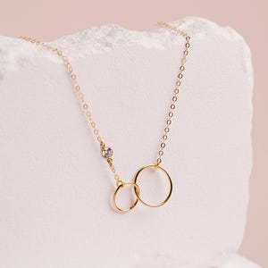 A delicate gold necklace with two smooth linked circles, one larger and one smaller, is displayed against a light pink background.