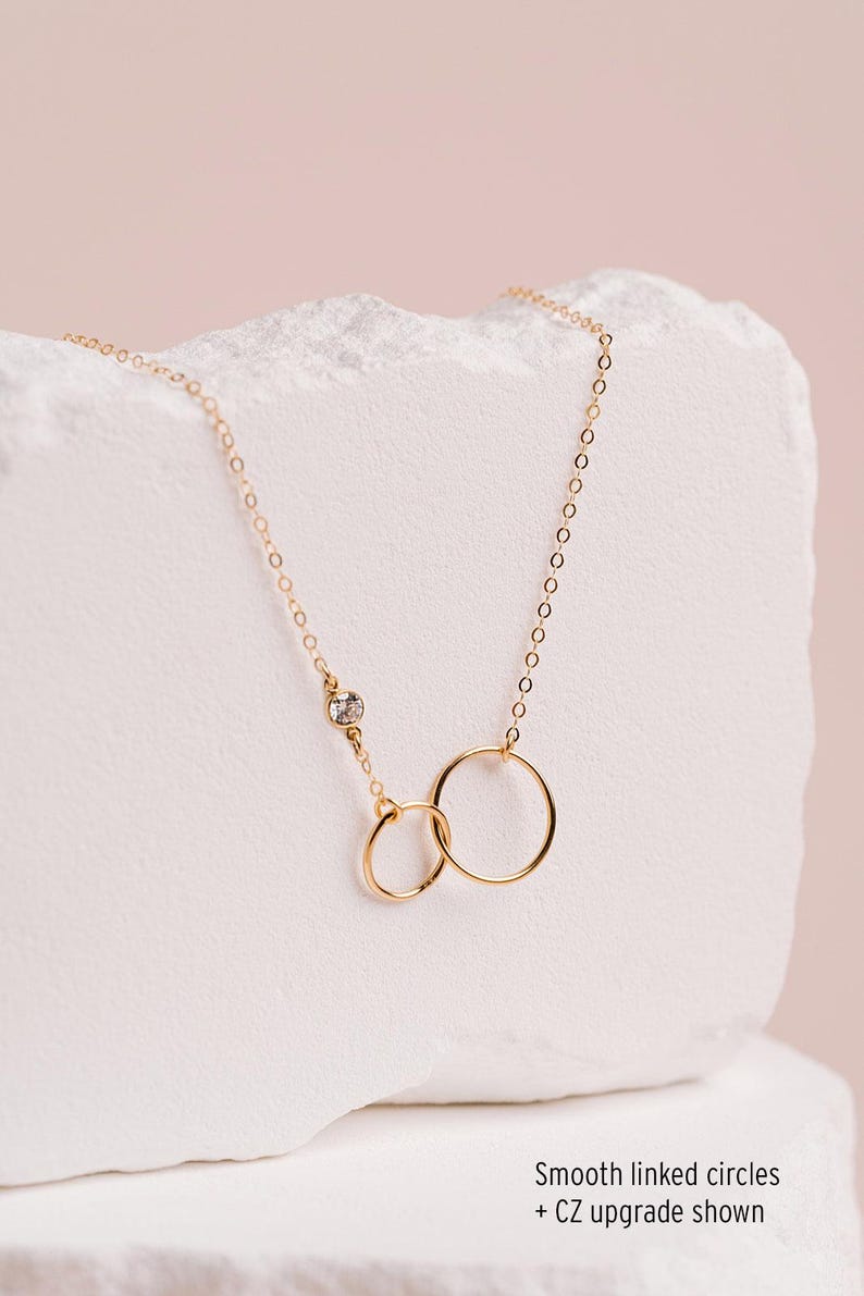 A delicate gold necklace with two smooth linked circles, one larger and one smaller, is displayed against a light pink background.