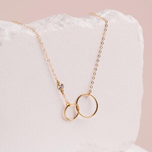 A delicate gold necklace with two smooth linked circles, one larger and one smaller, is displayed against a light pink background.