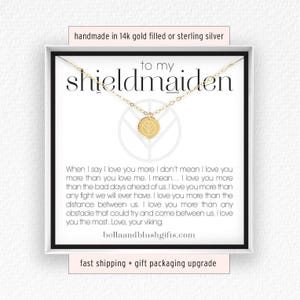 May include: A gold-toned necklace with a small circular pendant featuring a shield emblem, displayed on a white card with the text "to my shieldmaiden" and a long message about love, ending with "Love, your viking" and "bellaandblushgifts.com".