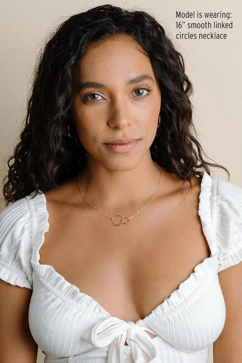 A young woman with dark curly hair wearing a white top and a necklace with a circular design.