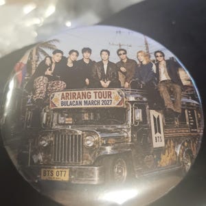 Pode incluir: Um bottom redondo com um grupo de sete pessoas sentadas em cima de um jeepney. O veículo exibe o texto ARIRANG TOUR BULACAN MARCH 2027, um logotipo do BTS e uma placa com a inscrição BTS OT7. O design apresenta uma estética vintage.
