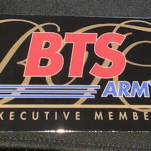 May include: A rectangular black card featuring the red text BTS above the blue text ARMY, underlined by three horizontal blue lines. The words EXECUTIVE MEMBER are printed in white at the bottom, with gold swirling line accents.