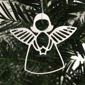 Pode incluir: Adorno de anjo de metal branco com asas e uma estrela no centro, pendurado em um galho de pinheiro verde.