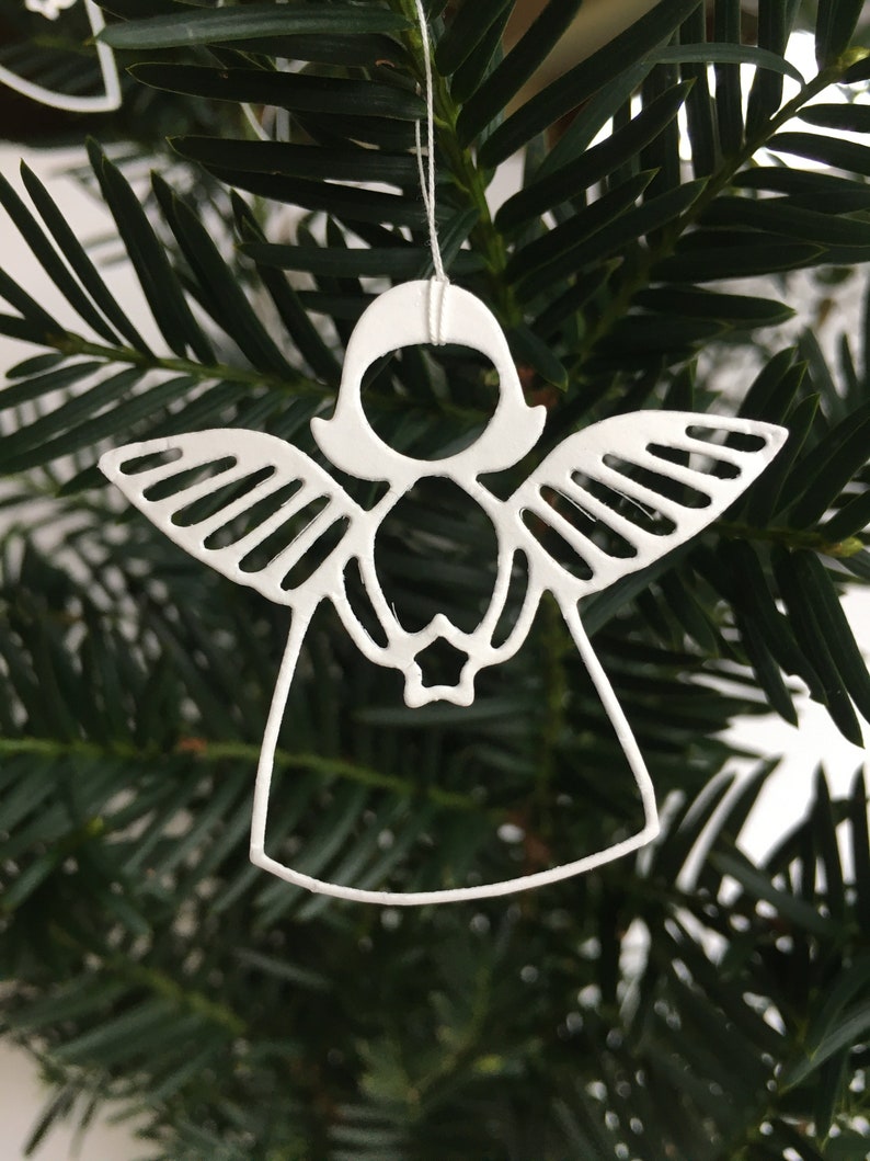 Pode incluir: Adorno de anjo de metal branco com asas e uma estrela no centro, pendurado em um galho de pinheiro verde.