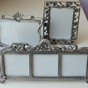 May include: A collection of three silver-toned metal picture frames. One frame is rectangular and vertical with floral detailing, one is horizontal with ornate scrollwork, and one is a long horizontal triple-pane frame with decorative borders.