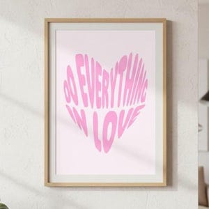 May include: A framed art print featuring a pink heart with the words "DO EVERYTHING IN LOVE" in a wavy font. The artwork is set against a light pink background and framed in a light wood frame.