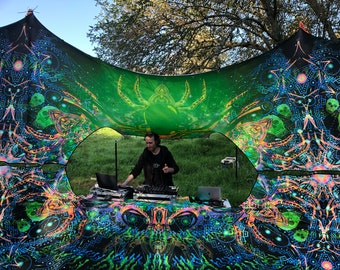 May include: A DJ set up outdoors under a colorful canopy with psychedelic patterns. The DJ is wearing a black shirt and is mixing music on a laptop and mixer.