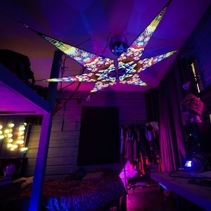 May include: A colorful, psychedelic star-shaped tapestry hangs from the ceiling of a room with a bed, a desk, and a clothing rack. The tapestry is made of fabric and has a variety of colors, including red, blue, green, and yellow. The room is lit with purple and blue lights.