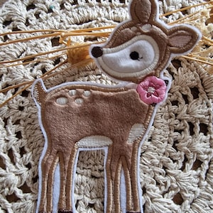 Könnte beinhalten: Ein braun-weißes Hirsch-Applikation mit einer rosa Blume und weißem Umriss. Das Reh hat einen weichen, strukturierten Körper und ist vor einem beige gehäkelten Hintergrund platziert. Ideal für Bastelprojekte.