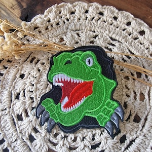 Könnte beinhalten: Gestickter Aufnäher mit einem grünen Tyrannosaurus Rex-Kopf mit offenem Maul, roter Zunge und weißen Zähnen. Der Aufnäher hat schwarze Umrisse und graue Krallen. Der Aufnäher liegt auf einer cremefarbenen Häkelspitze.