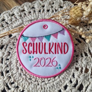 Könnte beinhalten: Ein runder, weißer, bestickter Aufnäher mit rosa Rand. Der Aufnäher zeigt das Wort "SCHULKIND" in rosa Schrift, darunter "2026". Ein Banner aus türkisfarbenen, rosa und lila Flaggen befindet sich über dem Wort.