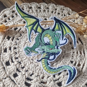 Könnte beinhalten: Ein gestickter Aufnäher, der einen grünen Drachen mit dunkelblauen Umrandungen und Akzenten zeigt. Der Drache hat große Flügel mit hellgrünen Innenteilen, einen langen, schlangenförmigen Körper mit gelbem Bauch, scharfe Krallen und ein rotes Auge.
