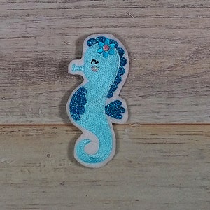 Könnte beinhalten: Ein hellblauer Glitzer-Seepferdchen mit einer blauen Blume auf dem Kopf. Das Seepferdchen hat ein lächelndes Gesicht und einen weißen Umriss.