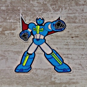 Könnte beinhalten: Ein gestickter Bügelaufkleber mit einem Roboter mit einem roten Umhang. Der Roboter ist blau und grün mit einem weißen Gesicht und steht mit ausgestreckten Armen.