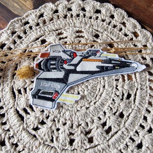 Könnte beinhalten: Gestickter Aufnäher mit einem Raumschiff-Design in Grau, Schwarz, Weiß, Rot und Gelb. Der Aufnäher trägt den Text "Star ship" und liegt auf einer beigefarbenen Häkelspitze.