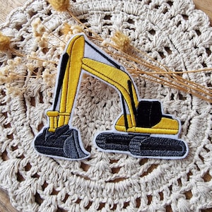 Könnte beinhalten: Gestickter Aufnäher eines gelb-schwarzen Baggers. Der Aufnäher zeigt ein detailliertes Design des Baufahrzeugs mit weißem Rand. Der Bagger ist vor einem neutralen Hintergrund platziert.