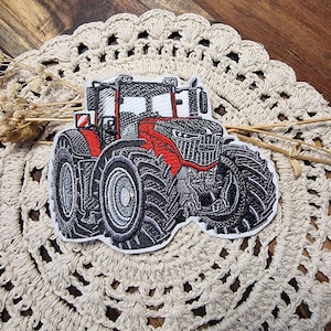 Könnte beinhalten: Gestickter Aufnäher mit einem detaillierten Traktormotiv. Der Traktor hat eine rote Motorhaube und einen schwarzen Körper mit großen, strukturierten Reifen. Der Aufnäher liegt auf einer cremefarbenen, gehäkelten Tischdecke.
