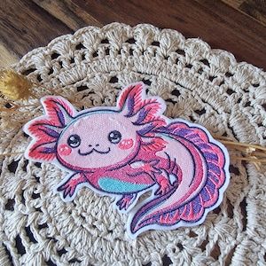 Könnte beinhalten: Gestickter Axolotl-Aufnäher mit einem rosa und lila Axolotl mit lächelndem Gesicht und detaillierten Kiemen. Der Aufnäher hat einen weißen Rand und liegt auf einer cremefarbenen Häkelspitze.