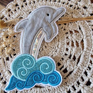 Könnte beinhalten: Ein grauer Delfin-Aufnäher mit weißem Rand, der über einer blau-türkisfarbenen Welle springt. Der Delfin hat einen schimmernden, irisierenden Akzent. Der Aufnäher ist für Kleidung oder Accessoires bestimmt.