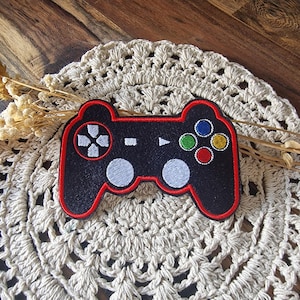 Könnte beinhalten: Ein schwarzer, gestickter Aufnäher mit einem Videospiel-Controller und einem roten Rand. Der Controller hat weiße, blaue, grüne, gelbe und rote Knöpfe. Der Aufnäher liegt auf einer cremefarbenen, gehäkelten Tischdecke.
