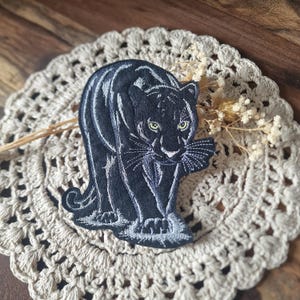 Könnte beinhalten: Ein gestickter Aufnäher, der einen schwarzen Panther in einer schleichenden Haltung zeigt. Das Design verwendet silber-grauen Faden für die Umrisse und Fell-Details sowie leuchtend gelb-grüne Augen. Der Panther ist von vorne abgebildet.