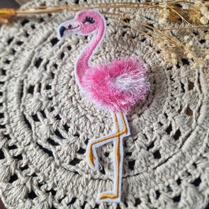 Könnte beinhalten: Ein pinkfarbener Flamingo-Aufnäher mit weißem Rand. Der Flamingo hat einen langen Hals, eine flauschige Brust und goldfarbene Beine. Der Aufnäher befindet sich auf einer beige gehäkelten Oberfläche.