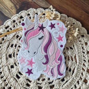 Könnte beinhalten: Ein gestickter Aufnäher mit einem weißen Einhornkopf und einer fließenden Mähne in Rosa- und Dunkellila-Tönen. Das Design enthält mehrere gestickte Sterne in Rosa und Dunkellila sowie kleine kreisförmige Akzente und geschwungene Muster.