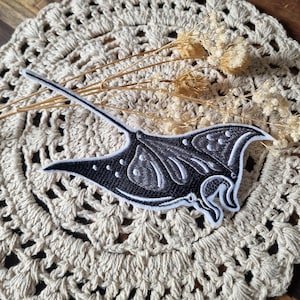 Könnte beinhalten: Gestickter Aufnäher mit einem schwarzen Mantarochen-Design und weißen Akzenten. Der Aufnäher hat ein detailliertes, gesticktes Aussehen und ist vor einem beigefarbenen, gehäkelten Hintergrund mit getrockneten floralen Elementen platziert.