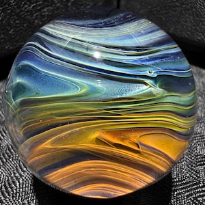 May include: A clear glass paperweight with a swirling design of blue, green, and orange colors. The colors blend together in a wave-like pattern, creating a unique and artistic appearance. The paperweight is round and has a smooth surface.