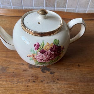 以下が含まれることがあります： ピンク、赤、黄色のバラの花柄が描かれた、金色の縁取りのある白いティーポット。ティーポットには、金色のつまみ付きの蓋が付いています。