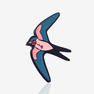 Puede incluir: Un pin con forma de golondrina con un diseño colorido. El pájaro tiene un cuerpo rosa, alas turquesas y un contorno negro. Sostiene una flor roja en el pico. El pin está sobre un fondo blanco.