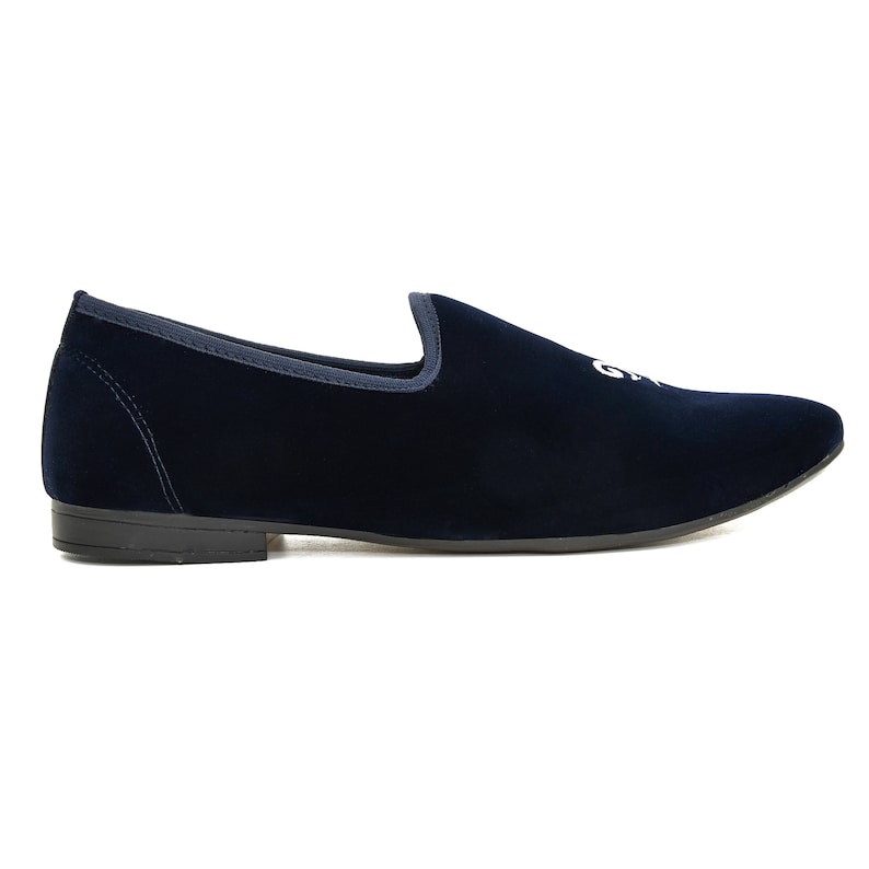 May include: A navy blue velvet slipper with a rounded toe and a low heel. The shoe features a white embroidered design on the upper. The shoe has a dark sole and a contrasting trim around the opening.