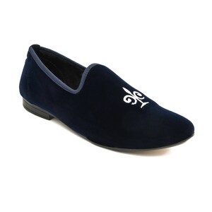 May include: A navy blue velvet slipper featuring a white embroidered fleur-de-lis. The shoe has a rounded toe and a dark blue trim. The sole is black.