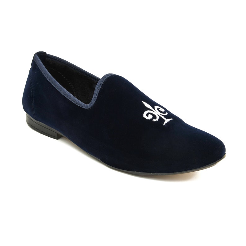 May include: A navy blue velvet slipper featuring a white embroidered fleur-de-lis. The shoe has a rounded toe and a dark blue trim. The sole is black.