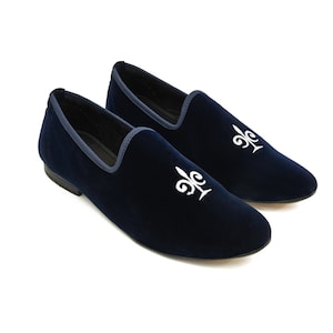 May include: Navy blue velvet loafers with white embroidered fleur-de-lis detailing. The shoes feature a dark blue trim and a low heel. These slip-on shoes are suitable for various occasions.